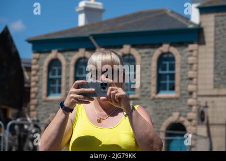 Ältere weibliche Touristin trägt Sonnenglas und ein gelbes Oberteil, das während des Urlaubs im Sommer Fotos auf einem Mobiltelefon fotografiert. Stockfoto