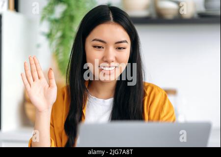 Positive glückliche hübsche chinesische junge Frau, die zu Hause in der Küche an einem Tisch mit einem Laptop sitzt, auf einer Online-Videokonferenz redet, winkt, mit Kollegen oder Freunden chattet, lächelt freundlich Stockfoto