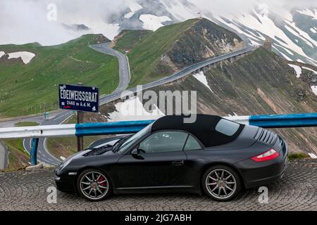 Porsche 911 Cabriolet mit geschlossener Oberseite steht vor dem offiziellen Hinweisschild Edelweissgrat 2460 Meter über dem Meeresspiegel auf Passstraße ab Stockfoto