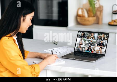 Online-Verhandlungen. Multirassische Gruppe von Menschen auf einem Laptop und eine asiatische Frau, die zu Hause in der Küche sitzt, sprechen auf einer Videokonferenz, entwickeln eine Projektstrategie, macht das Mädchen Notizen Stockfoto