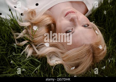 Glückliche blonde Frau, die sich auf dem grünen Gras legt und die Sommersaison genießt. Nahaufnahme Porträt von oben mit natürlichem Make-up Stockfoto