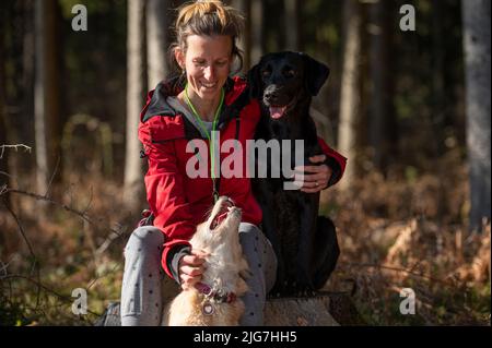 Junge Frau, die auf einem Baumstumpf in einem Wald mit ihren beiden Hunden neben ihr sitzt, in einem konzeptuellen Bild von Liebe und Vertrauen zwischen einer Besitzerin und ihrem Hund. Stockfoto