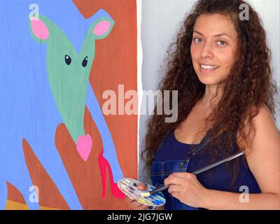Lächelnd schöne junge mexikanische Malerin (bildende Künstlerin) mit langen lockigen Haaren hält Pinsel und Palette vor ihrem Gemälde. Stockfoto