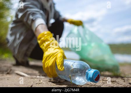 Eine junge Frau in gelben Handschuhen sammelt verlassenen Müll in einer schwarzen Tasche im Wald. Verschmutzung durch Plastik und Umweltschutz Stockfoto