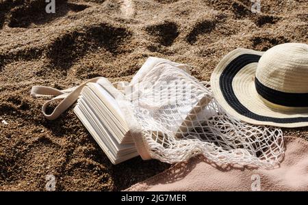 Strandzubehör: Umweltfreundliche Mesh-Einkaufstasche, Strohhut, Buch am Sandstrand Stockfoto