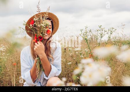 Porträt einer jungen Frau, die auf der Sommerwiese Wildblumen pflückt und nach Mohnblumen und Gänseblümchen riecht. Stockfoto