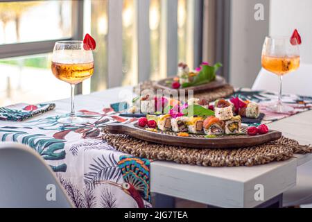 Eine Nahaufnahme von Sushi-Sets auf Tellern auf einem weißen Tisch mit einer tropischen Tischdecke und Tassen Champagner mit verschwommenem Hintergrund Stockfoto