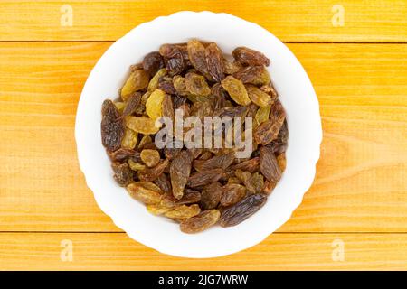 Rosinen in einer Schüssel. Dunkel getrocknete Weintraube auf einem dunkelweißen Teller auf einem orangefarbenen Holztisch. Goldene Rosinen. Braune und leichte Rosinen in einer Schüssel. Traditionell Stockfoto