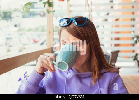 Junge Frau in violettem Hoodie mit blauem Becher, die auf der Terrasse heißen Tee oder Kaffee trinkt Stockfoto