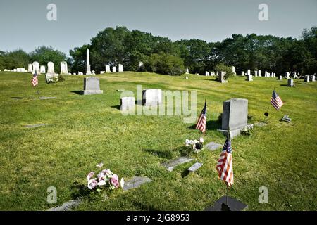 Weißer militärischer Grabstein an Grabstätten mit amerikanischen Flaggen. Militärfriedhof mit Reihen von Grabsteinen im Hintergrund Stockfoto