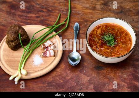 Ansicht von oben einer Schüssel Rote Beete Suppe mit Petersilie - ukrainischer Borscht - und einem Holzbrett mit grünen Zwiebeln, Dill, Salz, Speck und Vollkornrogen Stockfoto