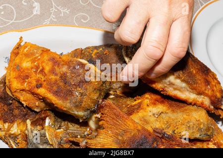 Beim Abendessen greift die Hand eines Mannes nach einem Stück gebratenem Karpfenfisch. Köstliche kalorienreiche Lebensmittel. Stockfoto