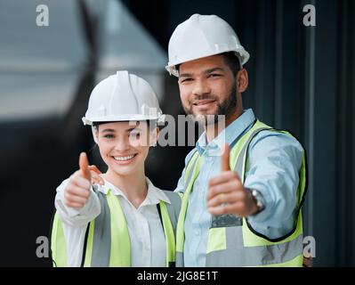 Ihre Baupläne wurden genehmigt. Beschnittenes Porträt von zwei jungen Bauarbeitern, die auf einer Baustelle den Daumen nach oben geben. Stockfoto