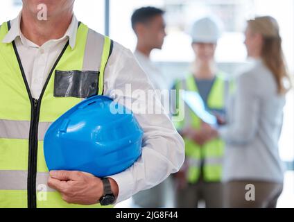 Lassen Sie uns an die Arbeit gehen. Ausgeschnittene Aufnahme eines nicht erkennbaren männlichen Bauarbeiters, der seinen Hardhut mit seinen Kollegen im Hintergrund hält. Stockfoto