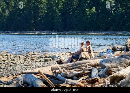 Ein Paar mit ihrem Hund sitzt auf Strandstühlen und genießt die Aussicht auf einen felsigen, holzbedeckten Strand an einem hellen Sommertag. Stockfoto