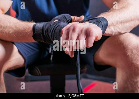 Kurzer Körper eines starken jungen Mixed-Kampfkünstlers, der auf der Couch sitzt und einen elastischen Verband einwickelt, um sein Handgelenk für einen offenen Faustkampf zu fixieren. Stockfoto