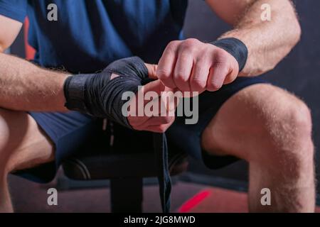 Kurzer Körper eines starken jungen Mixed-Kampfkünstlers, der auf der Couch sitzt und einen elastischen Verband einwickelt, um sein Handgelenk für einen offenen Faustkampf zu fixieren. Stockfoto