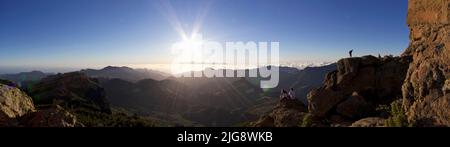 Spanien, Kanarische Inseln, Gran Canaria, Massif Central, Roque Nublo, Panoramaaufnahme, Sonnenuntergang, Sonne als Stern, blauer Himmel, Blick über das Massif Central im Westen, Teide, Teneriffa, Paar von hinten sitzend, zwei Personen neben dem Roque Stockfoto