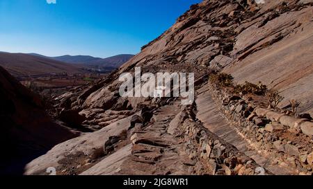 Spanien, Kanarische Inseln, Fuerteventura, Barranco de las Penitas, steiniger Weg am Rande des barranco zur Ermita de la Pena, blauer wolkenloser Himmel Stockfoto