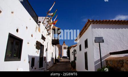 Spanien, Kanarische Inseln, Fuerteventura, Betancuria, alte Hauptstadt, Altstadt, Gasse, Kirche am Ende der Gasse, Fahnen, weiße Häuser, Holzbalkon, blauer Himmel, Verstreute weiße Wolken Stockfoto