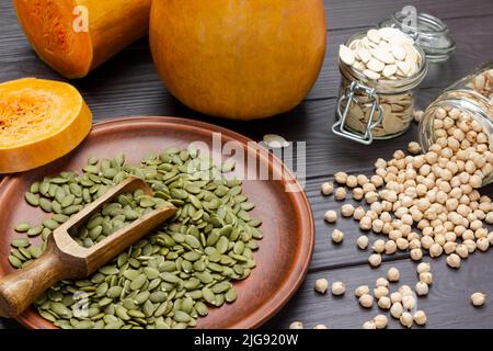 Geschälte Kürbiskerne auf dem Teller. Kichererbsen im Glasgefäß. Kürbis und Samen im Glas. Draufsicht. Dunkler Holzhintergrund. Stockfoto
