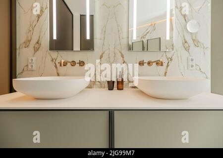 Vorderseite einer Toilette mit zwei Waschbecken mit rahmenlosen Spiegeln und goldenen Metallhähnen Stockfoto