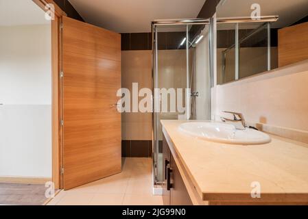 Badezimmer mit dunklen Holzmöbeln, cremefarbener Marmorplatte, rahmenlosem Spiegel und quadratischem Glas und einer Duschwanne aus Chrom-Metall Stockfoto