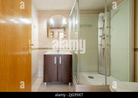 Badezimmer eines Hauses mit Duschkabine mit Massagedüsen, rahmenlosem Spiegel und Waschbecken aus dunklem Holz unten Stockfoto