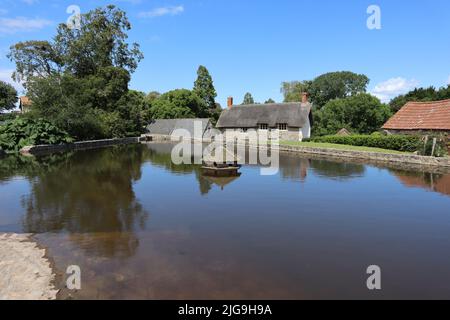 Das sechseckige Entenhaus auf dem Dorfteich bei East Quantoxhead in Somerset, England. Im Hintergrund sind reetgedeckte Hütten zu sehen Stockfoto