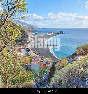 Eine Ozeanküste mit schwarzem Strandsand am Puerto de Tazacorte Strand von oben. Bunte Stadthäuser oder Ferienunterkünfte in der Nähe der Küste in Stockfoto