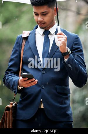 Ich kann alles passieren, egal was passiert. Ein junger Geschäftsmann, der ein Handy benutzt, während er an einem regnerischen Tag in der Stadt einen Regenschirm hält. Stockfoto