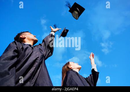 Würden es an die Spitze schaffen. Low-Angle-Aufnahme von zwei jungen Studenten, die ihre Hüte am Abschlusstag in die Luft werfen. Stockfoto