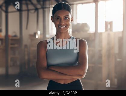 Seien Sie bereit, sich jeden Tag selbst herauszufordern. Porträt einer sportlichen jungen Frau, die mit gekreuzten Armen in einer Turnhalle steht. Stockfoto