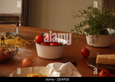 Ein Haufen frischer roter Tomaten in einer weißen Schüssel mit Eiern, Nudeln und Nudelmaschine auf einem Tisch Stockfoto