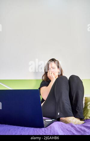 Eine vertikale Aufnahme einer kaukasischen Frau mit fokussierten Gesichtsaufnahmen, die vom Schlafzimmer aus auf den Computer blicken Stockfoto