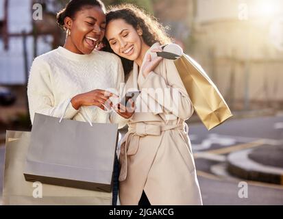 Kaufen Sie das Beste für viel weniger. Zwei junge Frauen, die ein Smartphone benutzen, während sie vor einem städtischen Hintergrund einkaufen. Stockfoto