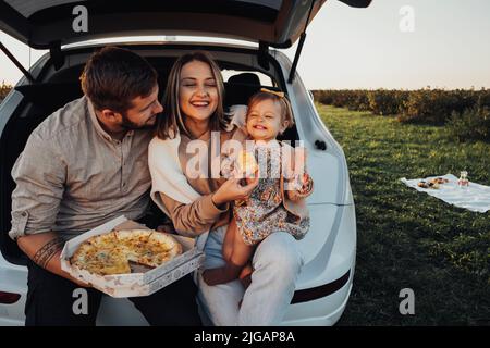 Fröhliche junge Familie, die im Kofferraum des SUV-Autos sitzt und Pizza isst, Mama, Papa und kleine Tochter, die am Wochenende im Freien Spaß haben Stockfoto