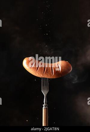 Einzelne leckere geröstete Grillwurst mit Gewürzen, die auf rustikaler Gabel vor dunkelschwarzem Hintergrund mit Dampf bestreut wurden. Kopierraum, vertikale Ausrichtung Stockfoto