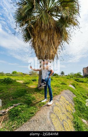 Eine vertikale Aufnahme eines schwulen Paares, das einen Videoanruf in der Nähe eines wachsenden Baumes macht Stockfoto