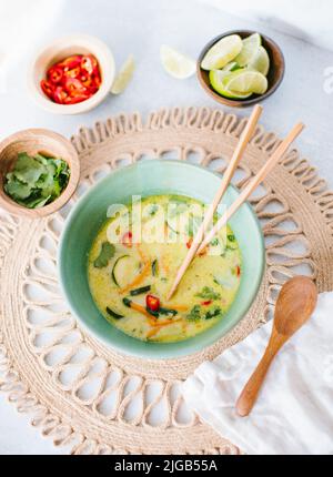 Eine vegane thailändische Kokosnusssuppe mit hölzernen Essstäbchen und einem Löffel, serviert mit würzigen Chilischoten, Koriander und Limette Stockfoto