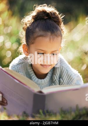 Ein kleines Mädchen, das ein Buch liest, während es sich in einem Park oder Garten entspannt. Gemischtes Rennen Kind lernen und bekommen eine Ausbildung, liegen auf dem Gras in der Natur und Stockfoto