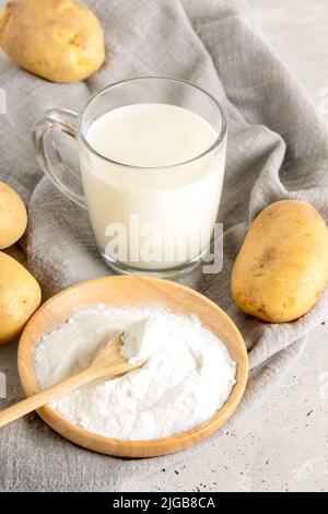 Gepulverte vegane Kartoffelmilch. Glasbecher mit Kartoffelmilch, Kartoffelpulver in Holzplatte und Knollen auf grauer Serviette auf Beton. Laktosefreies Gemüse mi Stockfoto