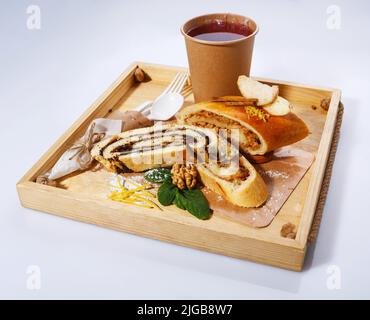Mohn- und Apfelstrudel auf einem rustikalen Holztablett auf weißem Hintergrund. Tee in einer Papiertasse und einem Löffel und einer Gabel aus umweltfreundlichen Materialien. Stockfoto