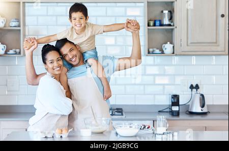 Portrait eines glücklichen jungen Mischling-Paares, das sich beim Backen mit ihrem entzückenden Sohn in einer heimischen Küche zusammenklebt. Hispanischer Ehemann, der ein Kind auf sich trägt Stockfoto