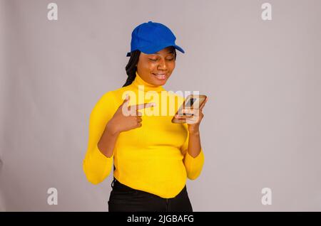Ein afrikanisches weibliches Modell, das auf ihr Smartphone zeigt, auf einem isolierten weißen Hintergrund Stockfoto
