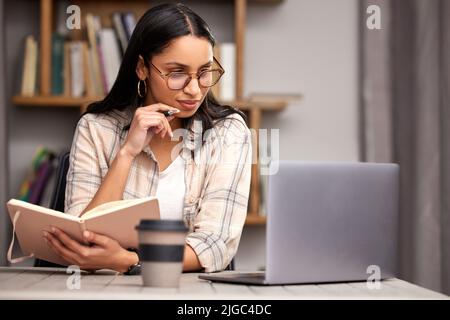 Fleißig in ihrem Studium. Eine attraktive junge Studentin, die sich Notizen macht, während sie mit ihrem Laptop in der Bibliothek studiert. Stockfoto