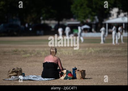 London, Großbritannien. 09.. Juli 2022. Eine Frau sitzt auf einem ausgetrocknten Rasen, während Menschen im Hintergrund Cricket spielen. Quelle: Sebastian Gollnow/dpa/Alamy Live News Stockfoto