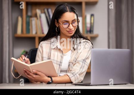 Sie weiß, wie man lernt. Eine attraktive junge Studentin, die sich Notizen macht, während sie mit ihrem Laptop in der Bibliothek studiert. Stockfoto