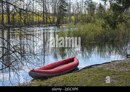 Rotes aufblasbares Kajak in der Nähe des Waldflusses. Frühlingswanderung zum Nationalpark. Stockfoto