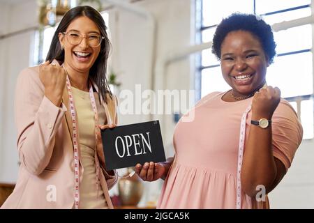 Wir sind offen für Geschäfte. Beschnittenes Porträt von zwei attraktiven jungen Geschäftsbesitzerinnen, die ein offenes Schild in ihrem Geschäft halten. Stockfoto
